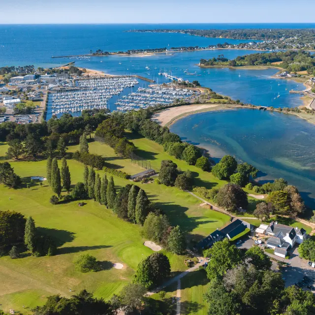 Foto, das eine Küste mit einer breiten Bucht/Mündung zeigt, einen Hafen mit vielen Booten auf der linken Seite, grüne Golfplätze im Vordergrund und Wohnhäuser auf der rechten Seite, dann das Meer im Hintergrund.