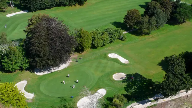 Luftbild eines Golfplatzes (grüne Rasenflächen, mehrere Bunker aus weißem Sand, Bäume, ein Gewässer unten rechts).
