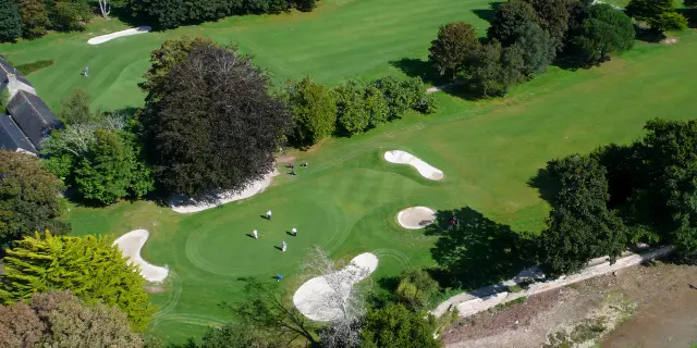 Luftbild eines Golfplatzes (grüne Rasenflächen, mehrere Bunker aus weißem Sand, Bäume, ein Gewässer unten rechts).