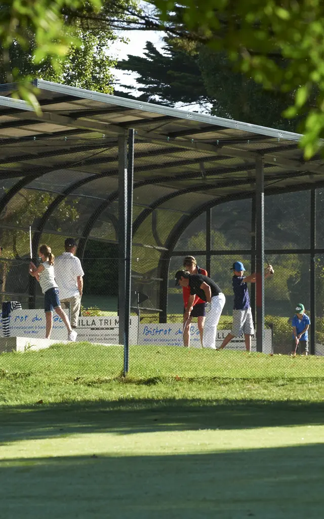 Une photo montrant un practice de golf: plusieurs personnes sous un long abri couvert, sur une pelouse, avec des arbres/feuillage au premier plan.