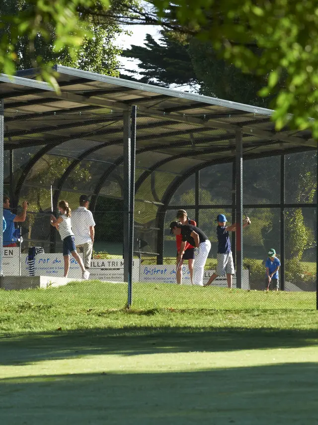 Une photo montrant un practice de golf: plusieurs personnes sous un long abri couvert, sur une pelouse, avec des arbres/feuillage au premier plan.