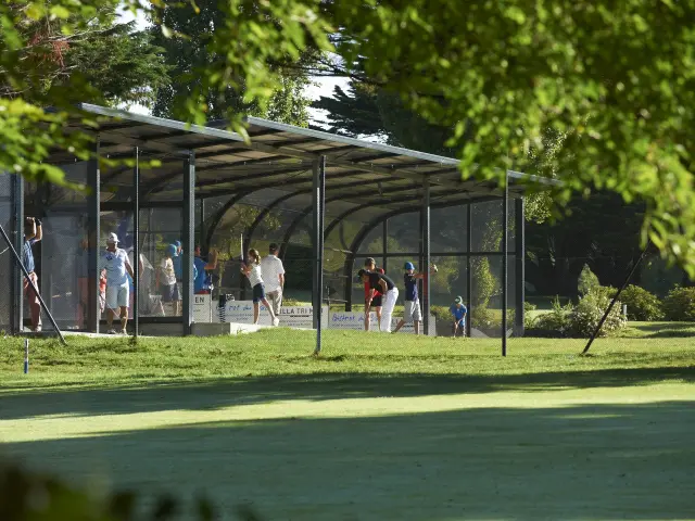 Une photo montrant un practice de golf: plusieurs personnes sous un long abri couvert, sur une pelouse, avec des arbres/feuillage au premier plan.