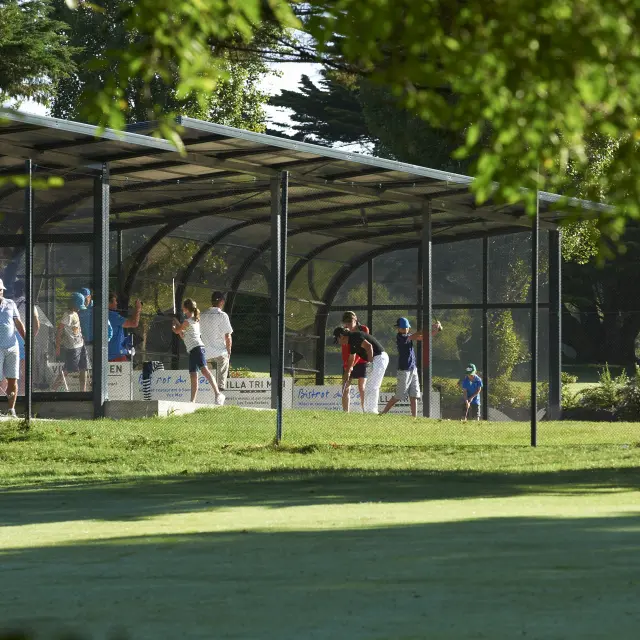 Une photo montrant un practice de golf: plusieurs personnes sous un long abri couvert, sur une pelouse, avec des arbres/feuillage au premier plan.