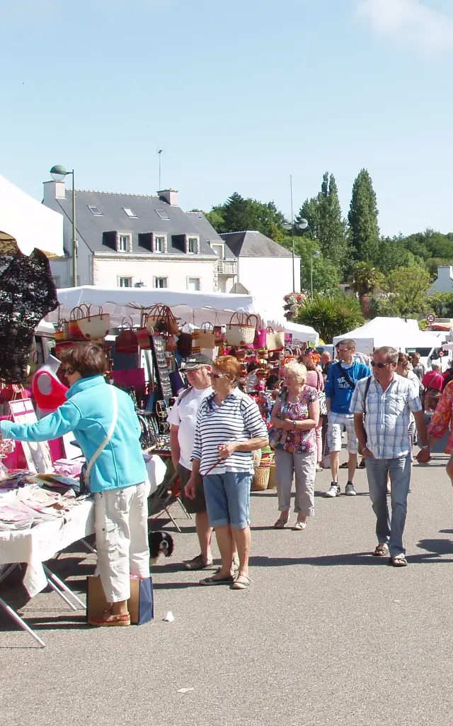 Une photo d’un marché en plein air. La photo montre une allée entre des stands sous tentes blanches, avec beaucoup de personnes qui circulent; au fond, des maisons et des arbres sous un ciel bleu.