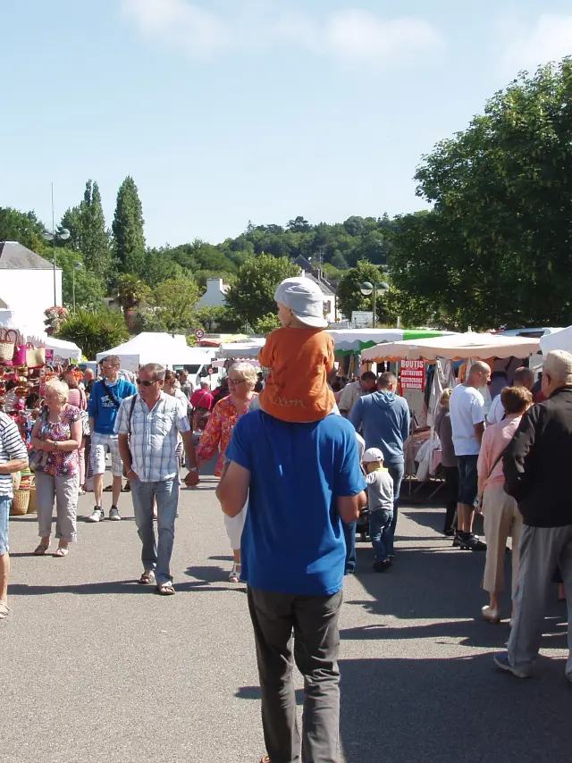 Een foto van een openluchtmarkt. Op de foto zie je een steegje tussen witte kraampjes met tenten, waar veel mensen rondlopen. Op de achtergrond zie je huizen en bomen onder een blauwe hemel.