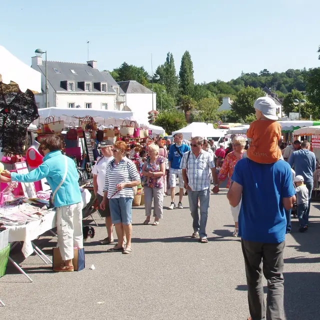 Une photo d’un marché en plein air. La photo montre une allée entre des stands sous tentes blanches, avec beaucoup de personnes qui circulent; au fond, des maisons et des arbres sous un ciel bleu.
