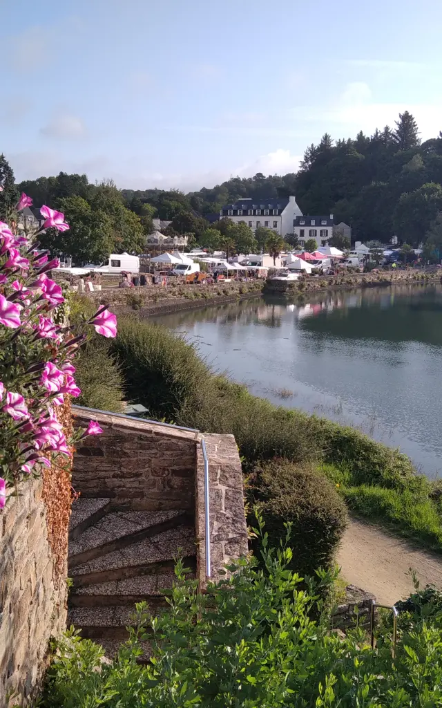 Une photo avec au premier plan à gauche, une grande masse de fleurs roses et blanches (type pétunias) qui débordent le long d’un muret en pierre. En dessous, un escalier en pierre descend vers un chemin. Au centre et à droite, un plan d’eau calme (rivière ou anse) longe une berge verdoyante avec un sentier. En face, des maisons blanches et des bâtiments sont visibles au bord de l’eau, et derrière eux une zone boisée. Le ciel est bleu clair.