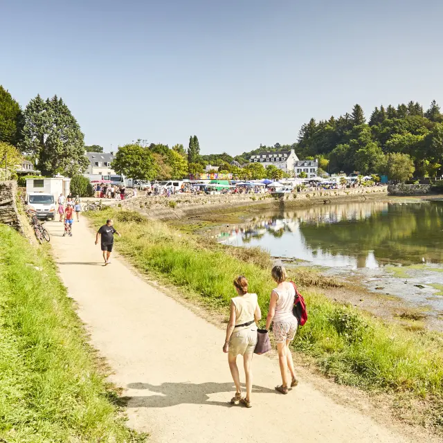 A photo of a footpath along an inlet at low tide, with grass at the edge; several people walking along the path; market stalls/vehicles in the distance; blue sky and trees on the opposite bank.