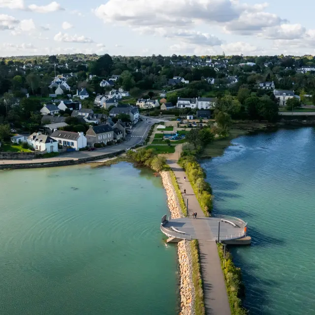 An aerial photograph shows a footbridge/river between two bodies of water, with a small circular widening in the middle; in the background, a village with houses and trees under a cloudy sky.