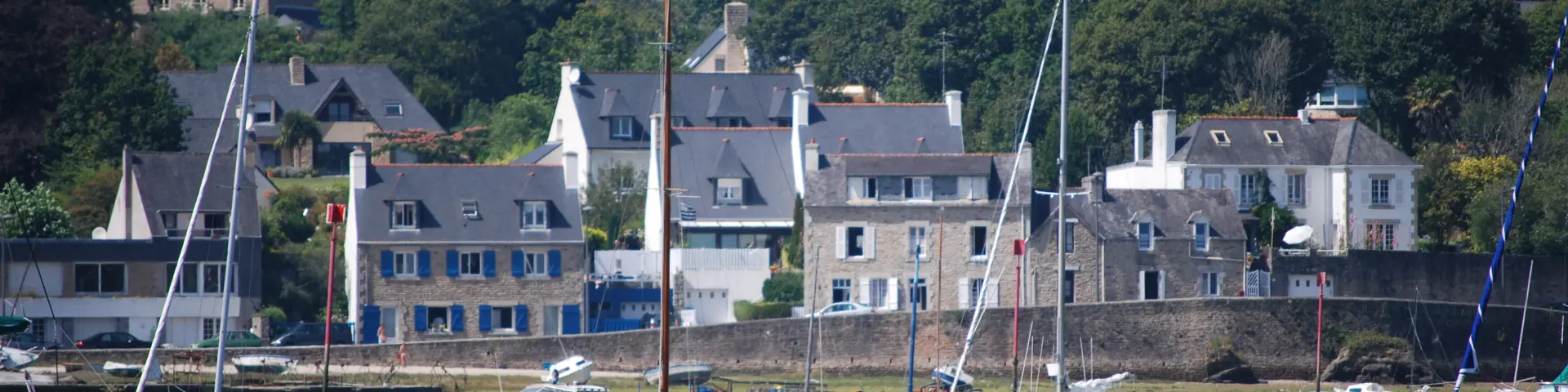 Das Foto zeigt einen Hafen mit blauem Wasser im Vordergrund, mehrere vor Anker liegende Boote (Segelboote und kleine Boote), weiße Bojen, dann Häuser aus Stein und mit blauen Fensterläden hinter einer Mauer, mit einem bewaldeten Hügel im Hintergrund unter blauem Himmel.