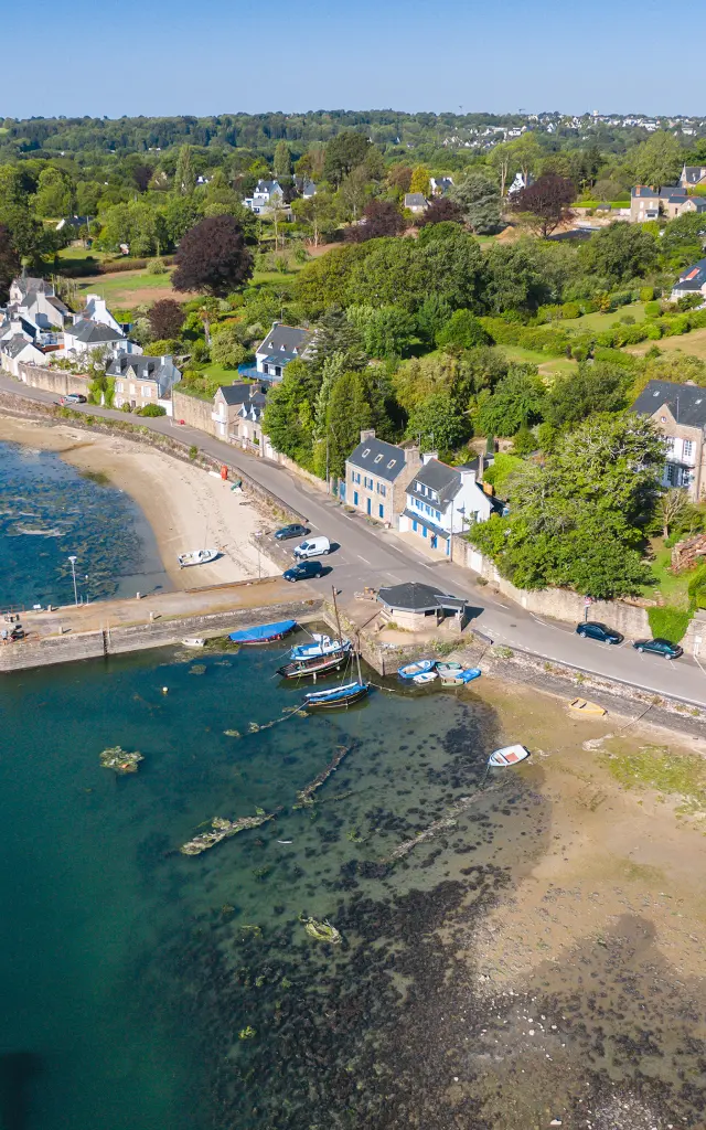photo aérienne d’un petit port en bord de mer. L’image montre une anse avec de l’eau bleu-vert, une plage, une petite jetée et quelques bateaux amarrés. À droite de la photo, une route longe des maisons et la végétation, sous un ciel bleu
