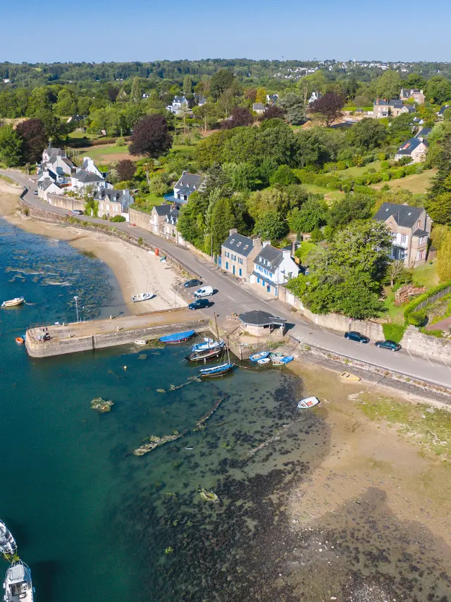 Luftbild eines kleinen Hafens am Meer. Das Bild zeigt eine Bucht mit blaugrünem Wasser, einen Strand, einen kleinen Steg und einige festgemachte Boote. Rechts im Bild führt eine Straße unter blauem Himmel an Häusern und Vegetation vorbei.