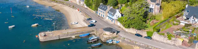 Luftbild eines kleinen Hafens am Meer. Das Bild zeigt eine Bucht mit blaugrünem Wasser, einen Strand, einen kleinen Steg und einige festgemachte Boote. Rechts im Bild führt eine Straße unter blauem Himmel an Häusern und Vegetation vorbei.