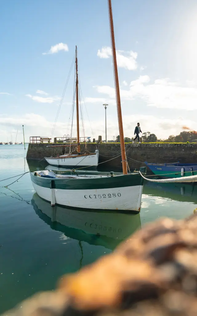 Das Foto zeigt einen kleinen, ruhigen Hafen: Ein Segelboot liegt am Kai, sehr glattes grün/blaues Wasser mit Spiegelungen, blauer Himmel mit tief stehender Sonne und einigen Wolken; im Vordergrund eine verschwommene Kaibegrenzung mit einigen Blättern. Masten und eine Straßenlaterne sind zu sehen, ebenso wie Gebäude und Autos im Hintergrund rechts.