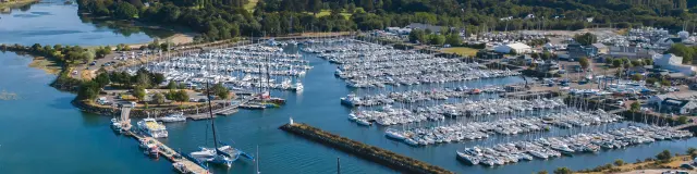 Een luchtfoto van een jachthaven met rijen afgemeerde boten, pontons, een golfbreker en een stuk blauwgroen water; in de verte zijn huizen en groene zones te zien.