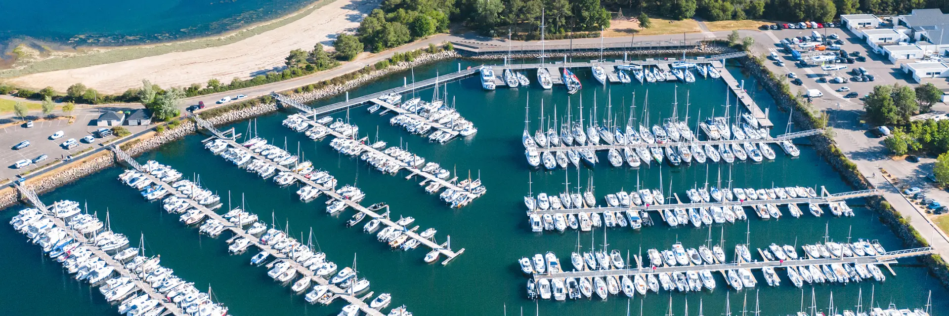 Luchtopname van een jachthaven vol jachten op pontons, met een strand en inham op de achtergrond en groene gebieden rondom.