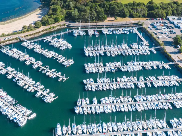 Luftbild eines Jachthafens voller Segelboote, die auf Pontons aufgereiht sind, mit einem Strand und einer Bucht im Hintergrund und Grünflächen drumherum.