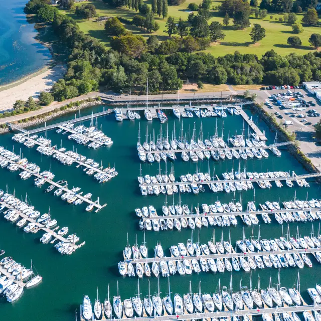Luchtopname van een jachthaven vol jachten op pontons, met een strand en inham op de achtergrond en groene gebieden rondom.