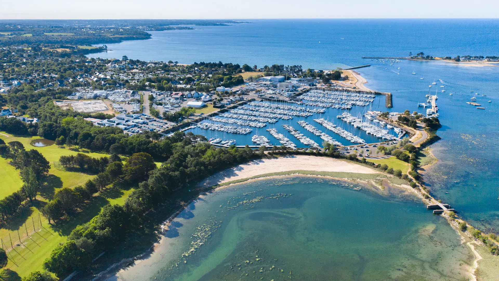 Luftbild eines Jachthafens am Meer. Das Foto zeigt einen mit vielen Booten gefüllten Yachthafen, Stege, einen Deich und Strände/Buchten mit türkisfarbenem Wasser sowie eine Grünfläche mit Bäumen im Vordergrund.
