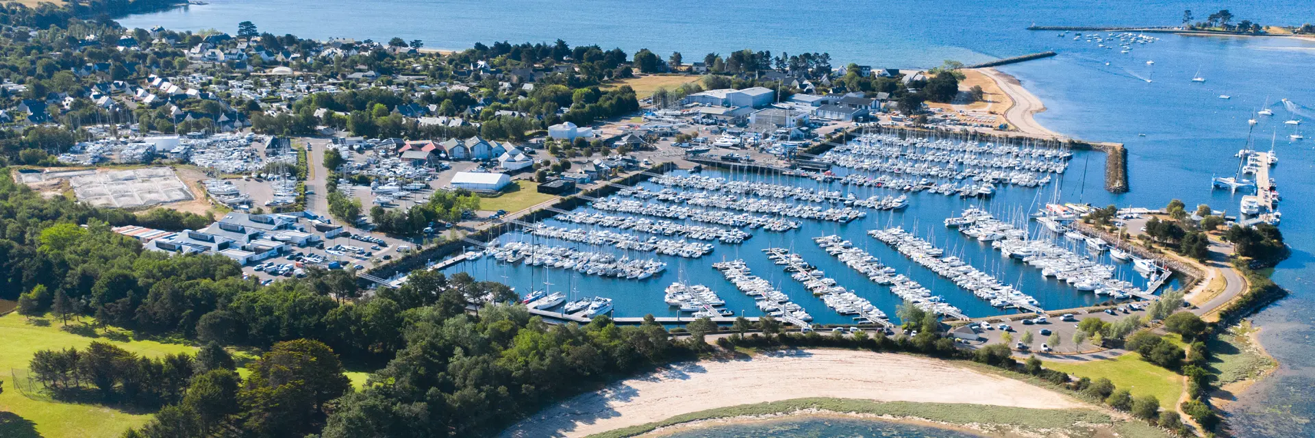 Aerial shot of a marina by the sea. The photo shows a marina full of boats, pontoons, a breakwater and beaches/beaches with turquoise water, and a green area planted with trees in the foreground.