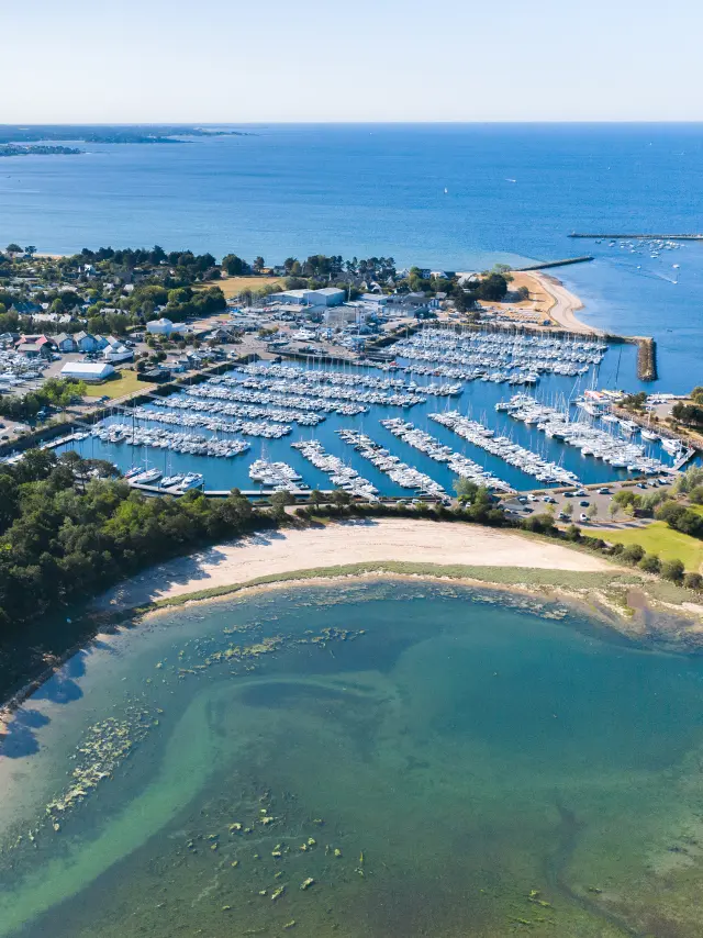 Aerial shot of a marina by the sea. The photo shows a marina full of boats, pontoons, a breakwater and beaches/beaches with turquoise water, and a green area planted with trees in the foreground.