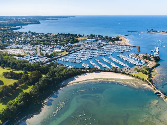 Aerial shot of a marina by the sea. The photo shows a marina full of boats, pontoons, a breakwater and beaches/beaches with turquoise water, and a green area planted with trees in the foreground.