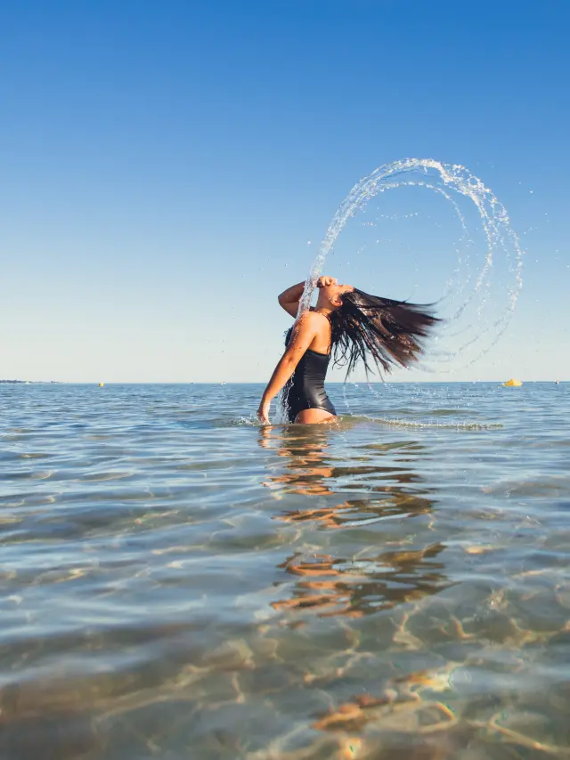 Ein Foto, das eine Person im flachen Wasser zeigt, die einen Badeanzug trägt und lange schwarze Haare hat, die nach hinten geworfen werden und einen Bogen aus Wasserspritzern bilden. Der Himmel ist blau, das Meer ruhig und der Horizont ist in der Ferne zu sehen.