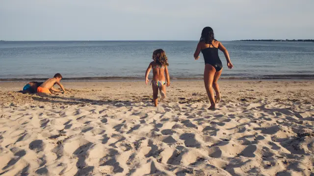 Ein Foto, das einen Sandstrand und das Meer am Horizont zeigt. Im Vordergrund sind drei Personen von hinten zu sehen: eine Erwachsene in einem schwarzen Einteiler in der Mitte rechts, die auf das Wasser zuläuft, ein Kind in der Mitte in einem bunten Zweiteiler, das ebenfalls auf das Wasser zuläuft, und ein weiteres Kind links, das im Sand liegt oder hockt (orangefarbenes Trikot). Klarer Himmel, ruhiges Meer.