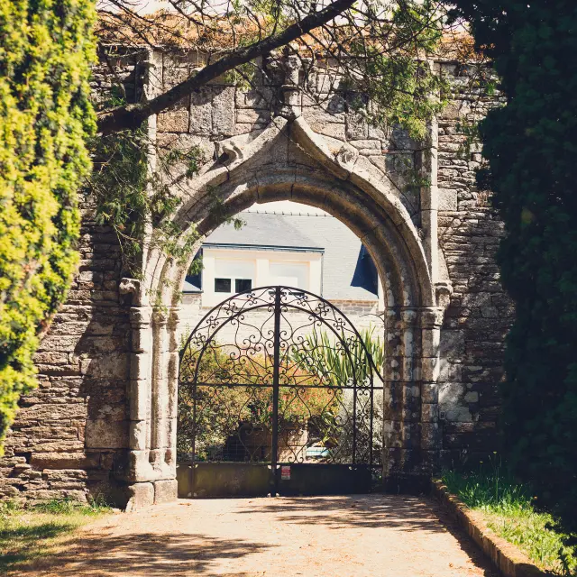 Ein Foto von einem Portal aus Stein. Das Tor ist ein großer gotischer Bogen aus grauem Stein mit einem geschlossenen schmiedeeisernen Gitter. Ein unbefestigter Weg führt zum Torbogen; Bäume/Äste rahmen die Oberseite und die Seiten ein, mit schattigen Bereichen auf dem Boden. Durch das Tor ist ein helles Gebäude mit Fenstern teilweise sichtbar.