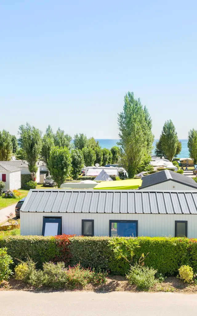 Ein Foto eines Campingplatzes, das Mobilheime zeigt, die entlang von Wegen aufgereiht sind, geparkte Autos, Hecken und Blumenbeete, hohe Bäume und das Meer, das in der Ferne unter einem blauen Himmel zu sehen ist.