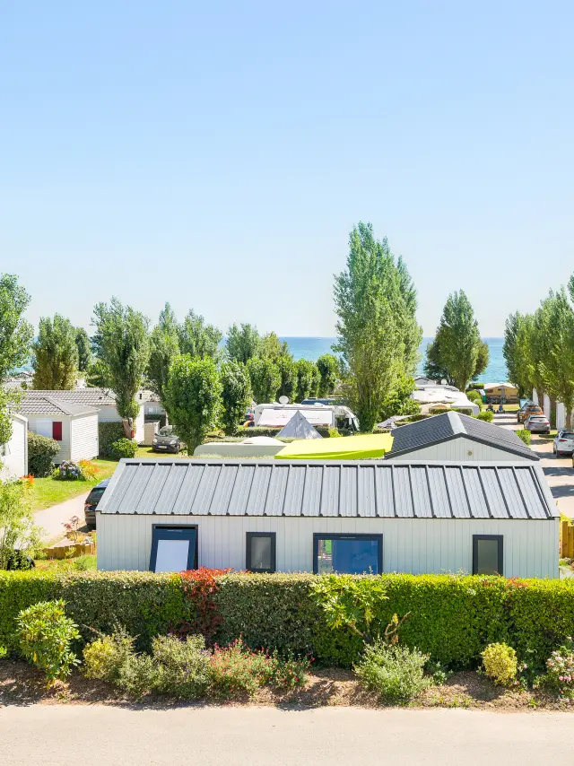 Une photo de camping qui montre des mobil-homes alignés le long d’allées, des voitures garées, des haies et des massifs de fleurs, des arbres hauts, et la mer visible au loin sous un ciel bleu.