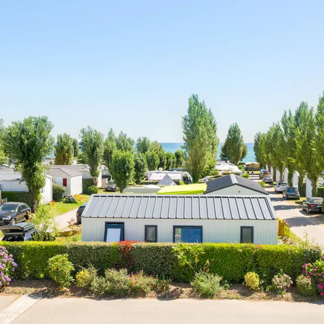 Une photo de camping qui montre des mobil-homes alignés le long d’allées, des voitures garées, des haies et des massifs de fleurs, des arbres hauts, et la mer visible au loin sous un ciel bleu.