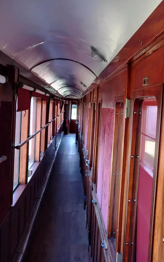 Ein Foto, das den Innengang eines alten Waggons zeigt, aus rotbraun lackiertem Holz, mit Fenstern auf der linken Seite, Türen/Kabinen auf der rechten Seite und einem Schild DUSCHEN mit der Nummer 7 darüber.
