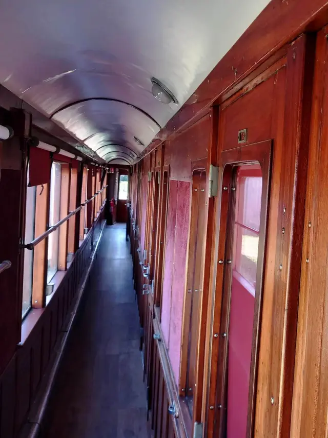 Une photo qui montre un couloir intérieur de wagon ancien, en bois verni rouge-brun, avec des fenêtres sur la gauche, des portes/cabines sur la droite et une plaque “DOUCHES” avec le numéro “7” au-dessus.