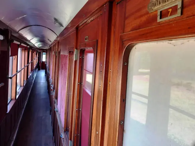 A photo showing an old wagon interior corridor, in red-brown varnished wood, with windows on the left, doors/cabins on the right and a SHOWER plaque with the number 7 above it.