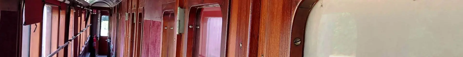 Ein Foto, das den Innengang eines alten Waggons zeigt, aus rotbraun lackiertem Holz, mit Fenstern auf der linken Seite, Türen/Kabinen auf der rechten Seite und einem Schild DUSCHEN mit der Nummer 7 darüber.