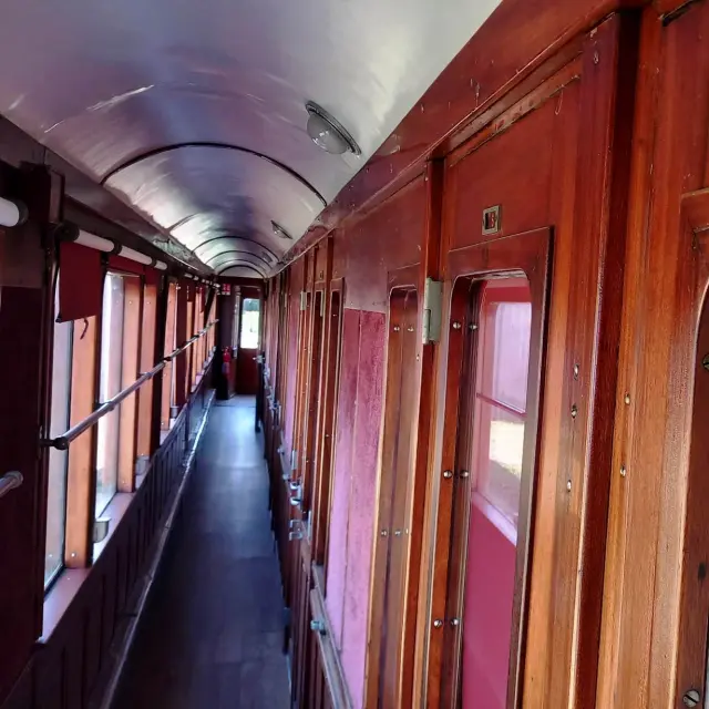 Ein Foto, das den Innengang eines alten Waggons zeigt, aus rotbraun lackiertem Holz, mit Fenstern auf der linken Seite, Türen/Kabinen auf der rechten Seite und einem Schild DUSCHEN mit der Nummer 7 darüber.