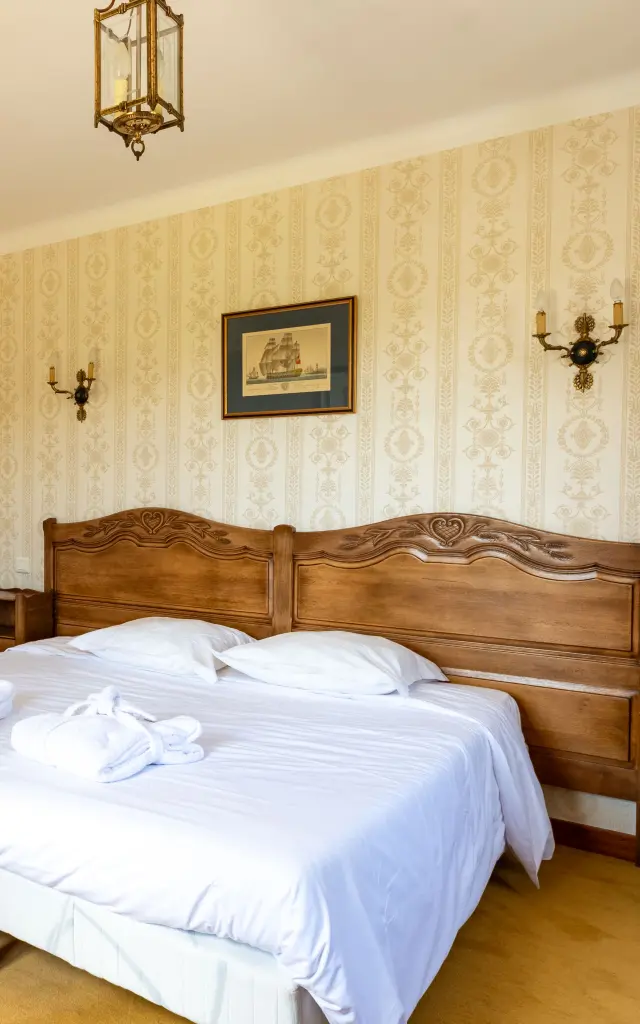 Ein Foto, das ein Hotelzimmer mit einem großen Doppelbett mit hölzernem Kopfteil, weißen Bettlaken und zwei zusammengerollten Handtüchern, die auf dem Bett liegen, zeigt. Beige gemusterte Tapete, zwei Wandleuchten, eine Deckenleuchte, ein Bilderrahmen über dem Bett. Links führt ein Bogen in einen anderen Teil des Raumes mit einem Wandspiegel und einem weiteren sichtbaren Bett im Hintergrund. Boden mit gelbem Teppich und Teppichen.