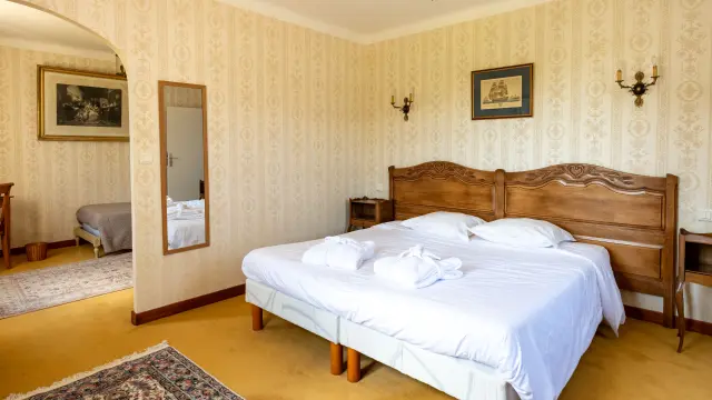 A photo showing a hotel room with a large double bed with a wooden headboard, white sheets and two rolled-up towels on the bed. Beige patterned wallpaper, two wall lights, a ceiling light and a picture frame above the bed. To the left, an archway leads to another part of the room with a wall mirror and another bed visible at the back. Floor with yellow carpet and rugs.