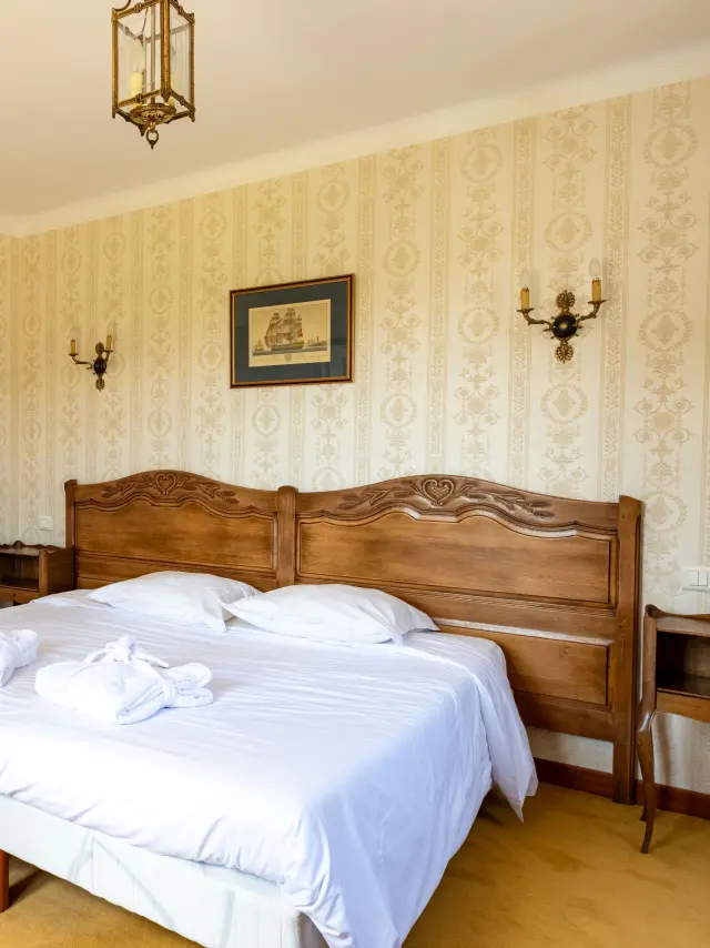 Une photo qui montre une chambre d’hôtel avec un grand lit double à tête de lit en bois, draps blancs et deux serviettes roulées posées sur le lit. Papier peint beige à motifs, deux appliques murales, un plafonnier, un cadre au-dessus du lit. À gauche, une arche mène vers une autre partie de la pièce avec un miroir mural et un autre lit visible au fond. Sol avec moquette jaune et tapis.