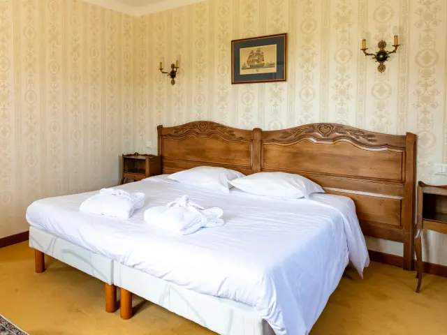 Une photo qui montre une chambre d’hôtel avec un grand lit double à tête de lit en bois, draps blancs et deux serviettes roulées posées sur le lit. Papier peint beige à motifs, deux appliques murales, un plafonnier, un cadre au-dessus du lit. À gauche, une arche mène vers une autre partie de la pièce avec un miroir mural et un autre lit visible au fond. Sol avec moquette jaune et tapis.