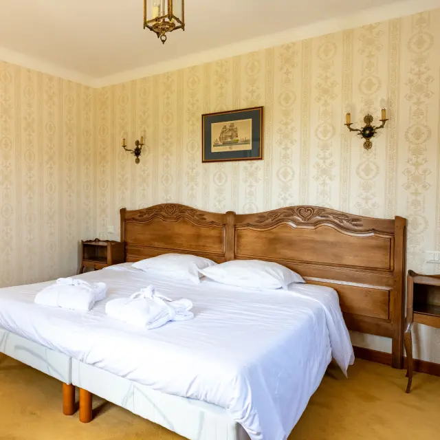 Une photo qui montre une chambre d’hôtel avec un grand lit double à tête de lit en bois, draps blancs et deux serviettes roulées posées sur le lit. Papier peint beige à motifs, deux appliques murales, un plafonnier, un cadre au-dessus du lit. À gauche, une arche mène vers une autre partie de la pièce avec un miroir mural et un autre lit visible au fond. Sol avec moquette jaune et tapis.
