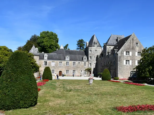 Das Foto zeigt ein großes Steingebäude im Stil eines Herrenhauses mit mehreren Schieferdächern, weißen Fenstern, einem hellen Hof/Terrasse davor und einem großen Garten im Vordergrund (Rasen, rote Blumenbeete, beschnittene Büsche). Klarer blauer Himmel.