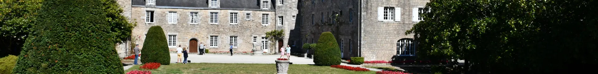 La photo montre un grand bâtiment en pierre de style manoir, avec plusieurs toits en ardoise, des fenêtres blanches, une cour/terrasse claire devant, et un grand jardin au premier plan (pelouse, massifs de fleurs rouges, buissons taillés). Ciel bleu dégagé.