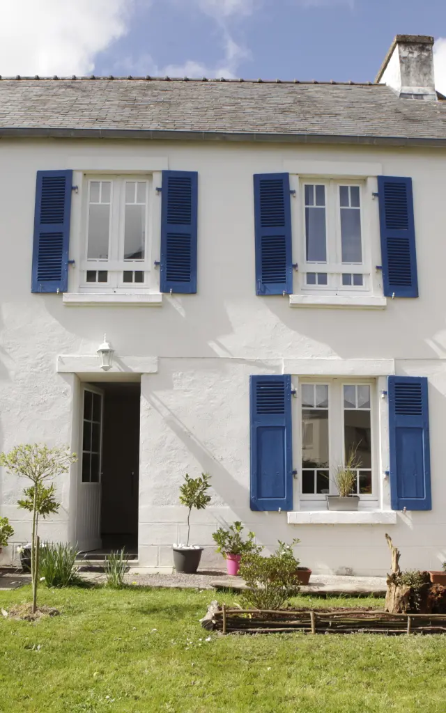 Ein Foto, das ein zweistöckiges weißes Haus mit blauen Fensterläden zeigt, eine Eingangstür in der Mitte, eine Terrasse mit schwarzem Tisch und Stühlen links, einen geschlossenen Sonnenschirm, ein Fahrrad, das rechts neben der Wand steht, und eine Rasenfläche im Vordergrund.