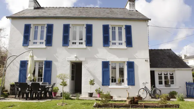 The photo shows a white two-storey house with blue shutters, a front door in the centre, a terrace with a black table and chairs on the left, a closed parasol, a bicycle next to the wall on the right, and a lawn in the foreground.