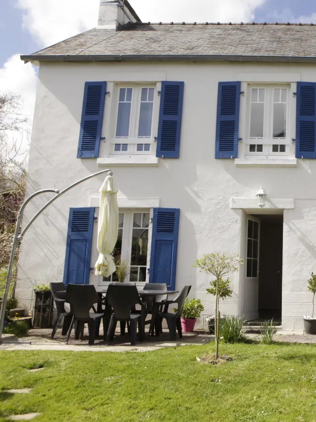 Une photo qui montre une maison blanche à deux étages avec volets bleus, une porte d’entrée au centre, une terrasse avec table et chaises noires à gauche, un parasol fermé, un vélo posé près du mur à droite, et une pelouse au premier plan.