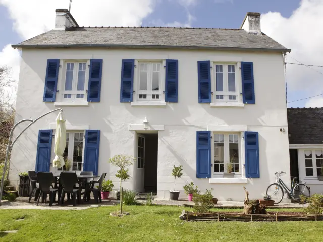 Une photo qui montre une maison blanche à deux étages avec volets bleus, une porte d’entrée au centre, une terrasse avec table et chaises noires à gauche, un parasol fermé, un vélo posé près du mur à droite, et une pelouse au premier plan.