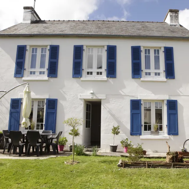 Une photo qui montre une maison blanche à deux étages avec volets bleus, une porte d’entrée au centre, une terrasse avec table et chaises noires à gauche, un parasol fermé, un vélo posé près du mur à droite, et une pelouse au premier plan.