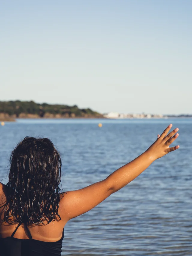 Une photo de mer qui montre une personne de dos, cheveux foncés mouillés, en maillot de bain noir, les deux bras levés en V face à la mer; horizon bas, ciel bleu clair, côte au loin.